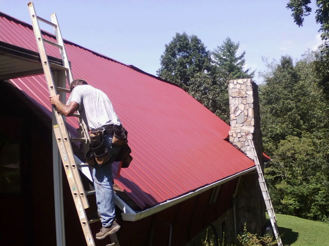 Roofer on a ladder beside a red metal roof and stone chimney — Soffit & Fascia Repair by Roofing Brothers in Stantonsburg